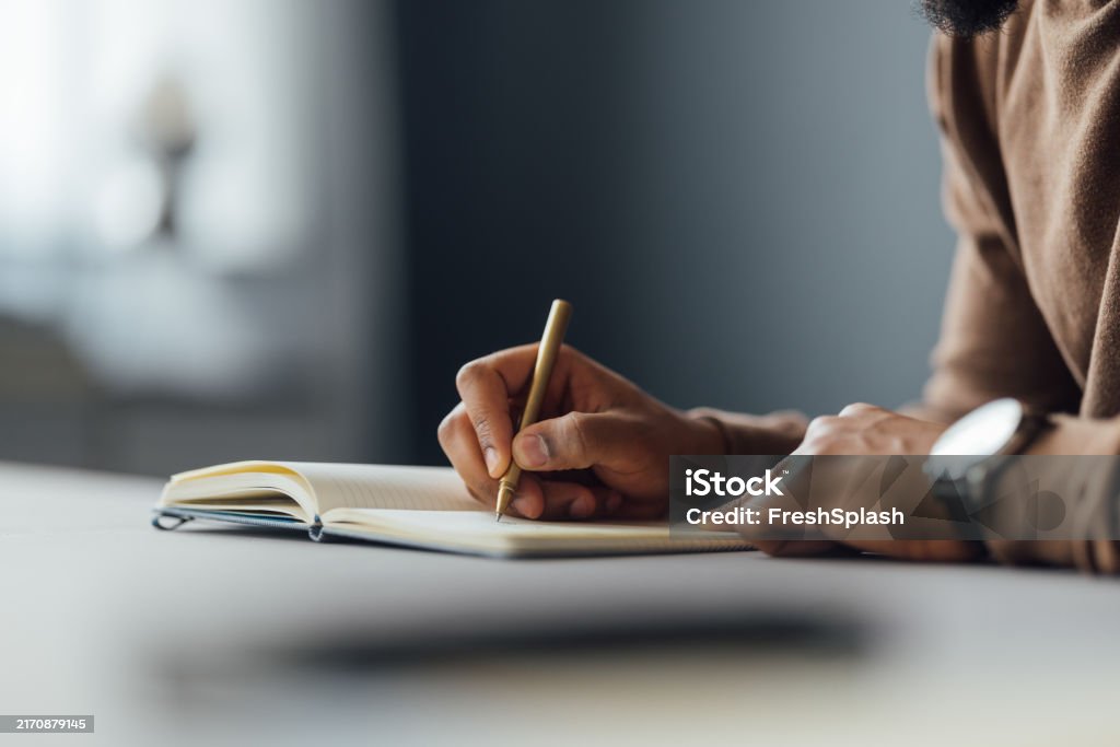 A close-up image of a man writing in a notebook with a pen at a desk, focusing on creativity and concentration.